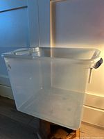 Clear plastic storage bin approximately 26 liters capacity, no lid visible, minor scuffs on surface, black side handles