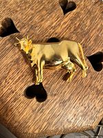 Front side view of small solid metal cow figurine with gold color, placed on wooden surface with heart-shaped cutouts.