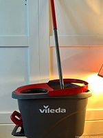 Side view of the Vileda brand spin mop bucket in red and gray plastic with the mop handle standing inside.