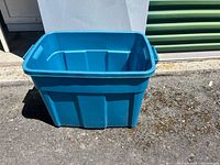 Blue plastic storage container placed outdoors on pavement, showing front and side view.