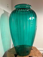 Front view of a large teal vintage glass vase with vertical ribbing, flared mouth, and rounded body, placed on a wooden surface against a white wall.