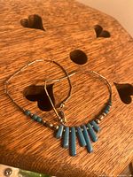 Photo shows a round wooden surface with heart-shaped cutouts, displaying a silver-tone hoop necklace with turquoise beads, with one broken elongated turquoise bead.