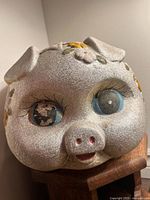 Front view of vintage metallic pig bank showing large blue eyes, one with worn paint, and painted yellow flowers on head.