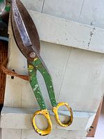Photo of a single pair of vintage metal tool scissors with yellow and green handles placed on a white surface.