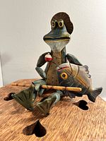 Front view of the wood carved frog figurine, showing the frog sitting with a carved fish and fishing pole in hand on a wooden surface.