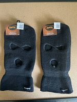 Two black Pioneer brand balaclavas laid flat showing eye and mouth holes and original tags attached.