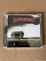Front cover of 'Fortress' album by Alter Bridge showing weathered house in field and album title.
