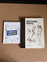 Front covers of the two books side by side on a brown surface showing 'How to Cartoon' and 'Sketching People' titles.