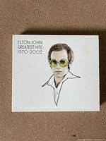 Front cover of the Elton John Greatest Hits 1970-2002 CD box set showing a stylized drawing of Elton John in yellow glasses.