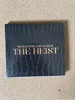 Front cover of the CD album The Heist by Macklemore & Ryan Lewis with black textured design and gold text.