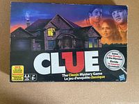 Photo of the Clue board game box showing the house illustration and characters on the front, with text about it being a classic mystery game and suitable for 2 players.