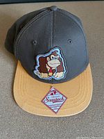 Front view of dark gray and mustard yellow snapback hat with embroidered Donkey Kong patch and 'Original Snapback' sticker on brim.