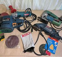 All four Bosch power tools with cords, sanding discs, dust bag visible
