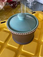 Closed view of the pottery jar showing the brown unglazed exterior with white wavy rim design and blue glazed lid with knob handle.
