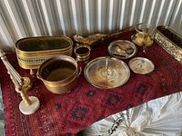 Photo showing a collection of brass items on a red patterned cloth, including a candle holder with marble base, planter, bowl, funnel, bird figurine, plates, and a creamer pitcher.