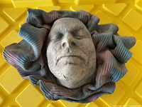 Close-up of front side of clay mask wall art showing closed-eye face and textured surface with ruffled multicolored border.