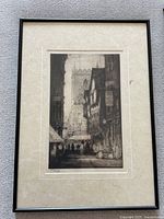 Framed black and white print showing a street scene with a church tower and old buildings, in a black frame with white matte.