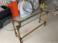 Rectangular clear glass top table with rounded long edges on a bronze/brass-colored metal frame with decorative legs. Shows the full table and the small chip on the glass.