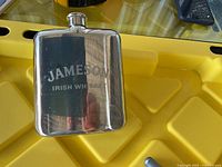 Stainless steel Jameson branded hip flask placed on a yellow surface, showing front view with reflective finish and screw cap at top.