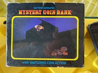 Photo of the front box showing the product name, image of the black coin bank with hand snatching coins, and the phrase 'with SNATCHING COIN ACTION'