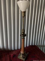 Full view of the tall brass and glass lamp with white conical shade against a corrugated metal background.