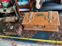 Image showing three decorative boxes of varying sizes and styles displayed on a patterned rug with a yellow tape measure indicating the largest box's length.
