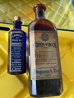 Photo showing two vintage apothecary bottles on a yellow tray, one blue labeled for croup and whooping cough embrocation and one brown labeled MORRH-VIN-OL.