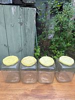 Four clear square glass jars with yellow lids on wooden surface