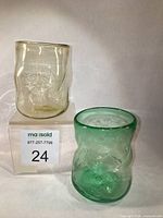 Two Ted Jolda blown glass tumblers, one green and one light amber, shown side by side on clear stands.