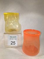 Two hand-blown glass tumblers, one yellow and one orange, placed on clear acrylic stands against white background. Both show organic shapes and smooth finishes.