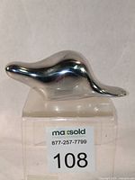 Front view of polished aluminum abstract beaver sculpture on clear base, showing smooth contours and label.