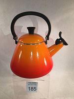 Front and side views of the Le Creuset volcanic orange whistling kettle with black foldable handle and spout cover.