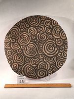 Full front view of the round ceramic wall hanging with spiral patterns and measurement ruler at base showing approx. 16 inches diameter.