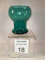 Front view of the teal crackle glass vase showing bulbous top and cylindrical base, placed on a clear acrylic display block with lot number sticker.