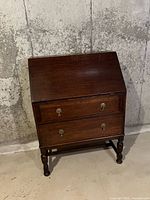 Front view of the antique dark brown wood secretary desk, closed, showing two drawers with metal handles and turned legs.