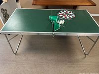 Full view of mini ping pong table with green tabletop, white lines, metal folding legs, green net, 4 white balls, dart board placed on table.