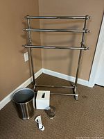 Photo of chrome towel rack with multiple bars, metal waste basket, white tissue box cover, small glass container, and white plastic object