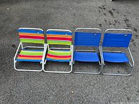 Four beach chairs placed side by side on asphalt, two with striped fabric and two with solid blue fabric seats and backs.