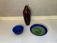 Lot overview showing the dark red ceramic vase and two glass bowls, one blue and green shallow bowl, and one cobalt blue signed bowl.