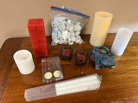 Group of various candles and candle holders on a wooden surface, including tall red square candle, battery powered candles, glass votives, frog shaped holder, and bag of tealight candles.