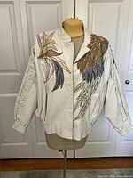 Front view of the creamy white leather jacket on a mannequin, showing detailed embroidered leaf patterns in beige, gold, blue, and black.
