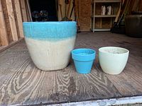 Photo of three ceramic planters on wood floor. Largest planter beige with turquoise top, medium planter fully turquoise, and smallest planter cream color.