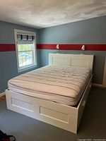 Full view of white queen size wooden bed frame with mattress on top, showing paneled headboard mounted on wall with two reading lamps attached.