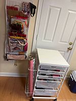 White metal frame craft cart with five mesh drawers filled with various craft supplies, standing on a wooden floor by a white door and wall-mounted gift bag organizer.