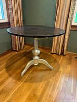 Full side view of the small wooden round table showing the dark stained table top and champagne pedestal base in a corner of a room with curtains and hardwood floor.