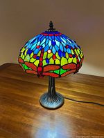 Front view of Tiffany style stained glass table lamp on wooden surface, lit to show multicolored stained glass shade with blue, red, green, yellow colors and electric cord visible.