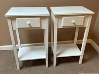 Photo showing front view of two white bedside tables with drawer and shelf; tables show signs of wear and surface marks.