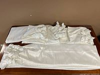 Four folded white silk-like curtain panels laid on a wooden surface, showing the smooth texture and length of the fabric.