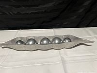 Full side view of the stainless steel pea pod art centerpiece showing five spheres inside the elongated pod shape on a white cloth with a black background.