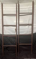 Photo showing two wooden blanket ladders standing upright against a white backdrop with a black base, clearly displaying their size and structure.